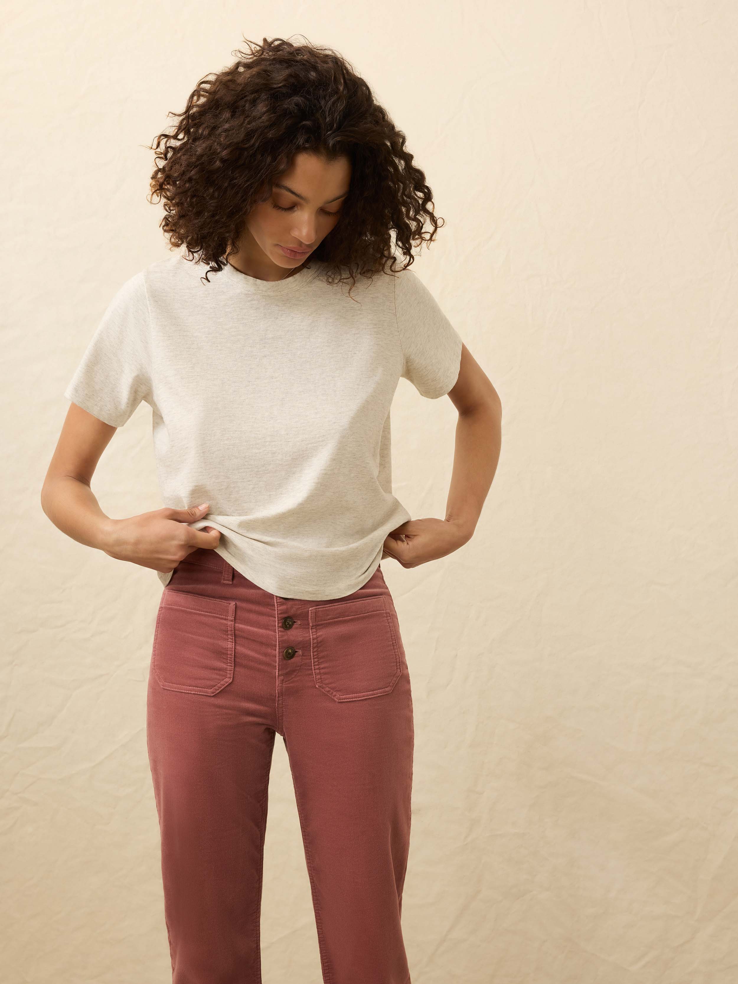 Woman wearing a light heather grey t-shirt and pink corduroy pants against a beige background