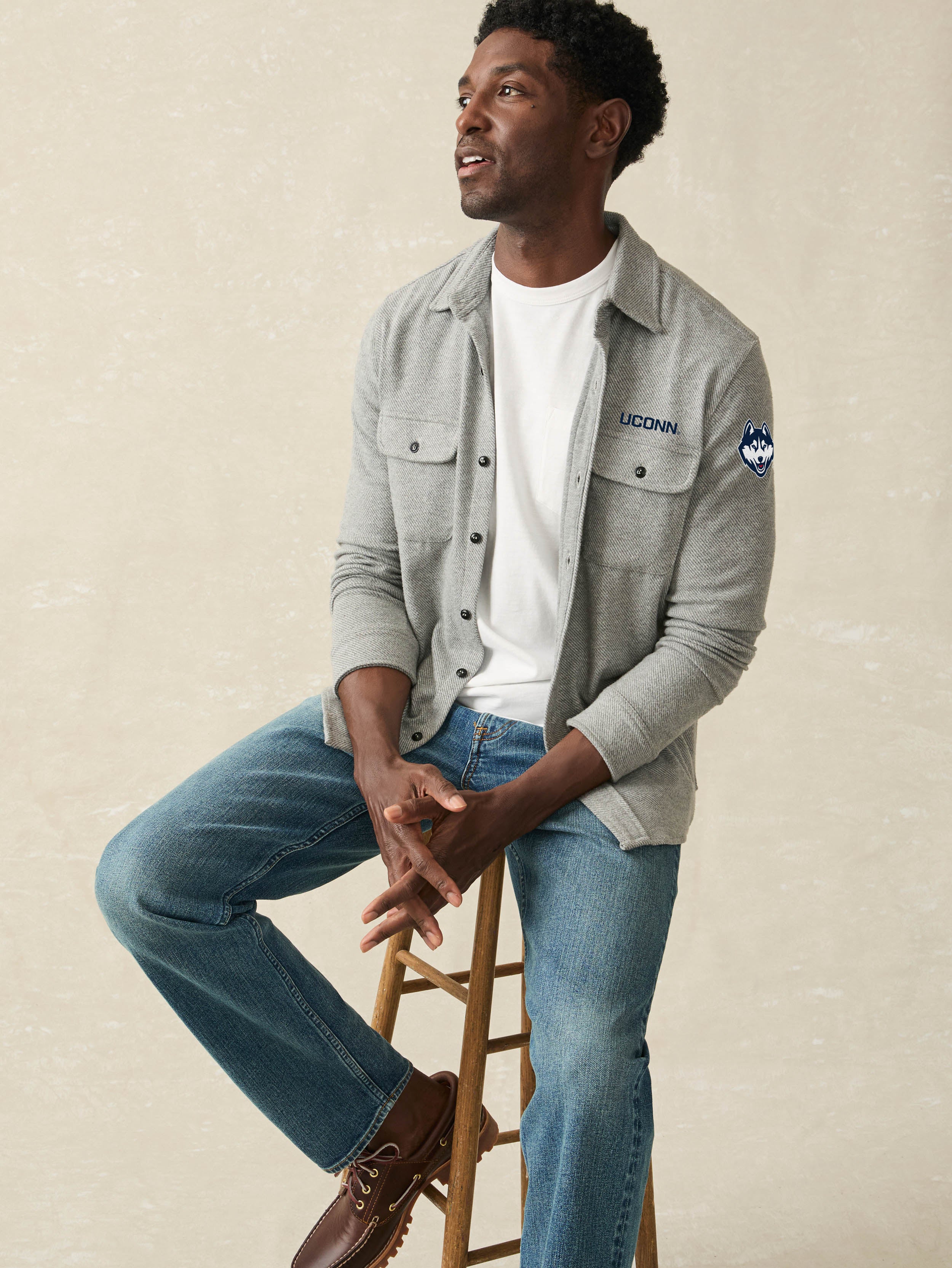 Man is wearing a grey long sleeve shirt with UCONN graphic on left chest and left arm against a beige background