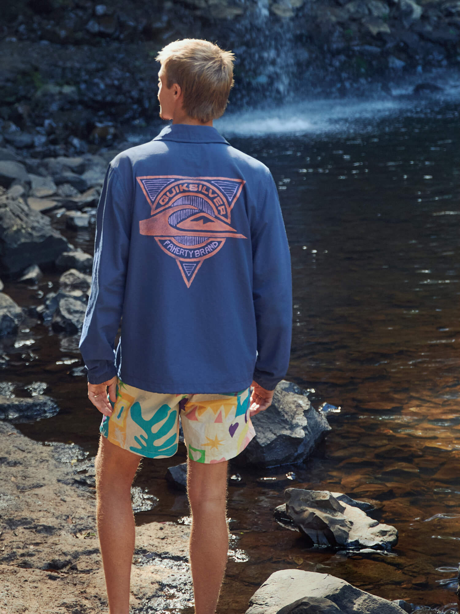 Man wearing a blue jacket with a Quiksilver x Faherty brand logo and colorful swim shorts standing by a waterfall.