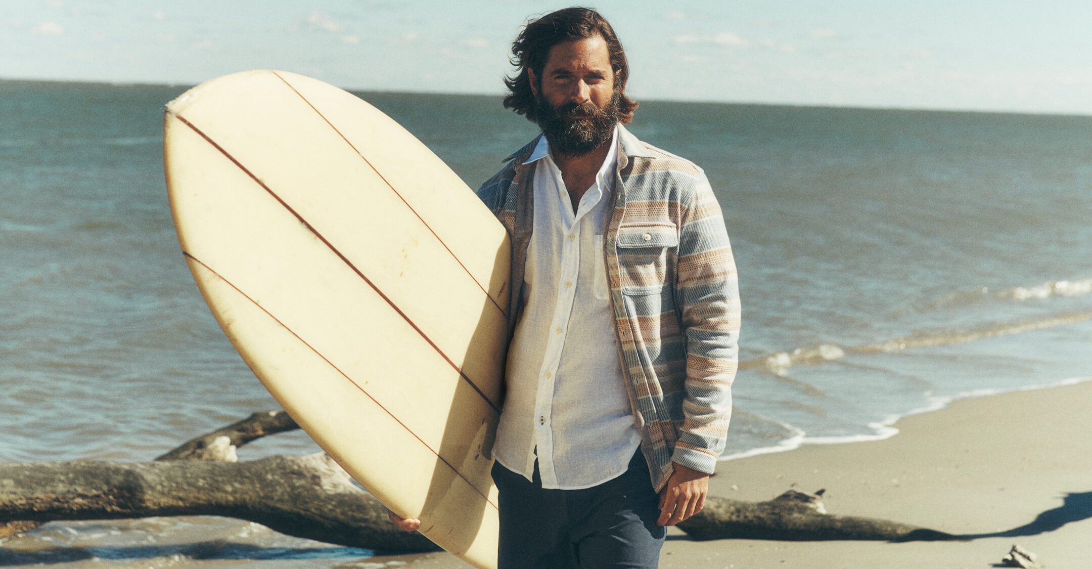 Man on the beach holding a surfboard wearing a legend sweater shirt over a short sleeve linen shirt paired with all day shorts