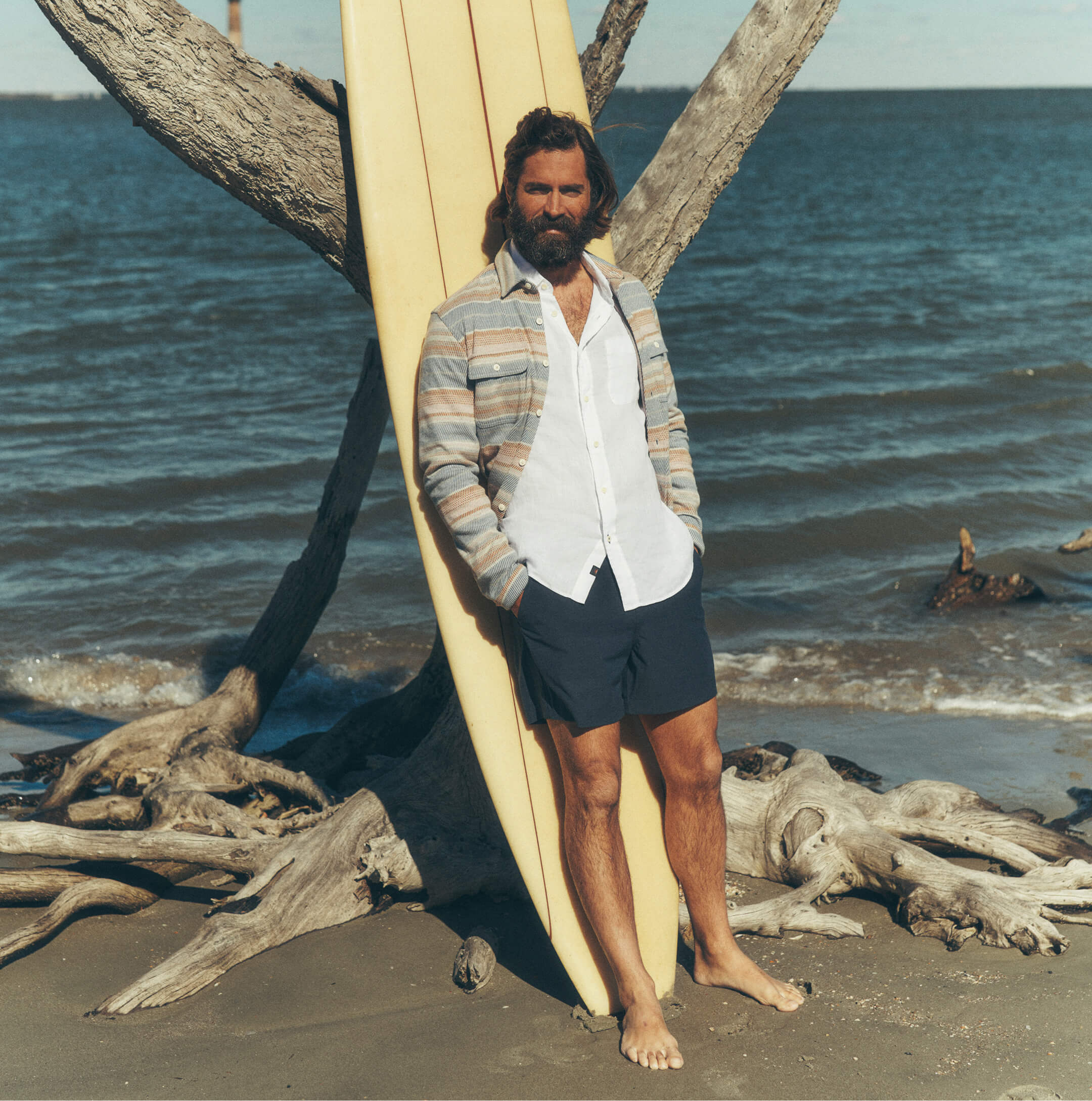 Man on the beach leaning against a surfboard wearing a legend sweater shirt over a short sleeve linen shirt paired with all day shorts