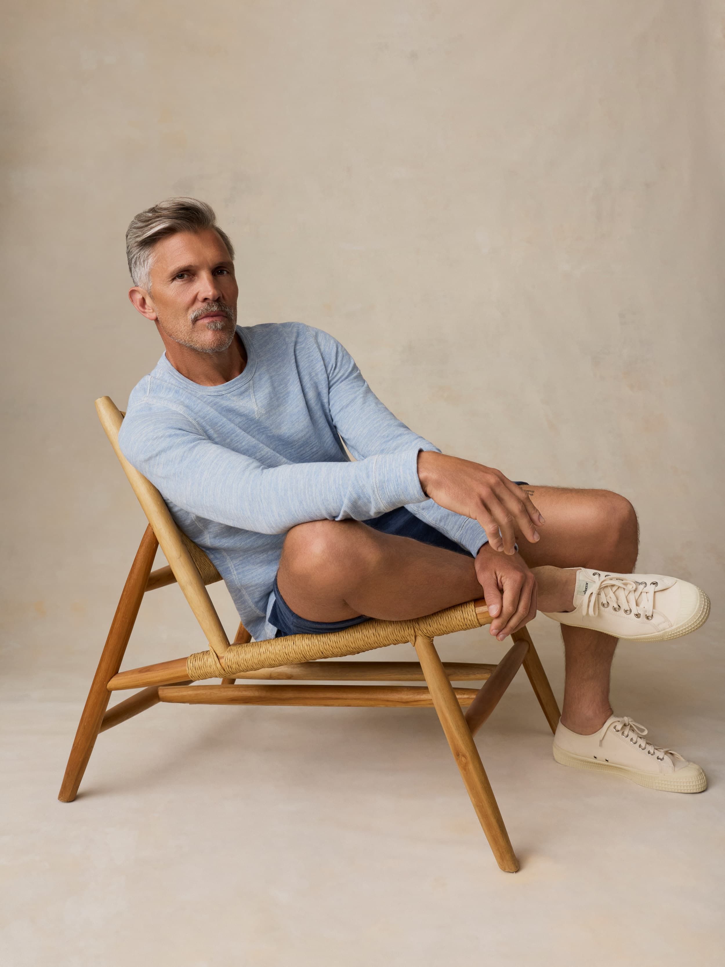 Man sitting on a wooden chair wearing a light blue crewneck and shorts against a beige background