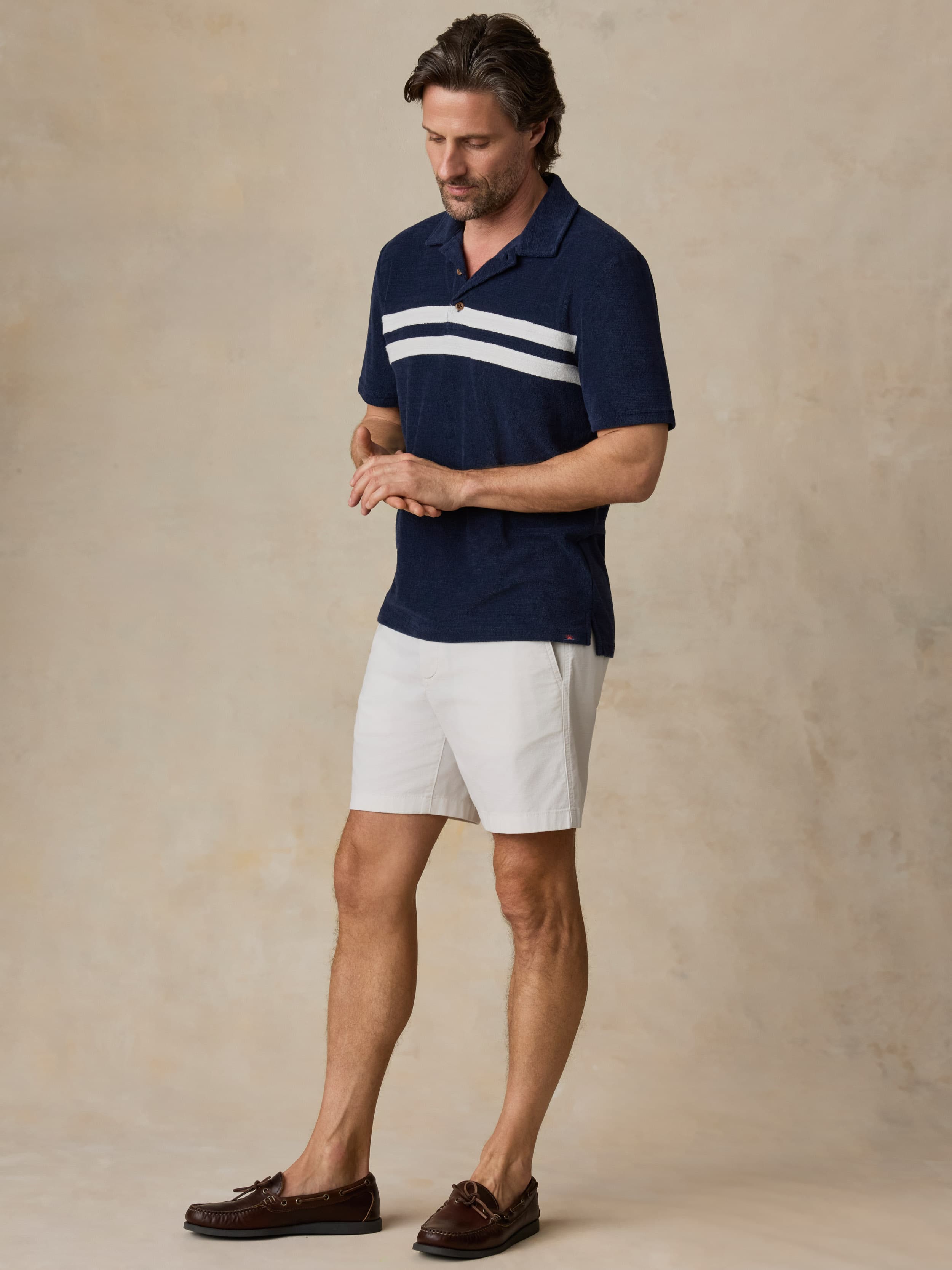 Man wearing a navy polo shirt with white stripes and white shorts against a beige background
