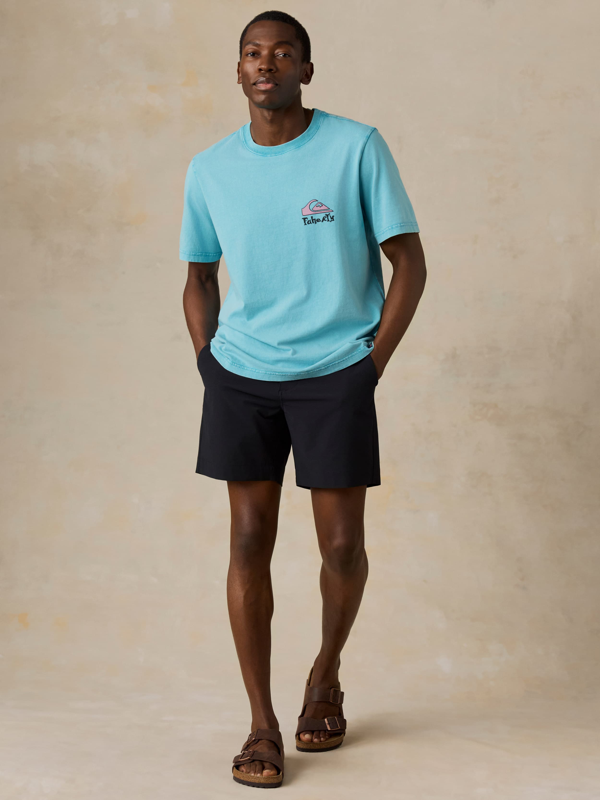 Man wearing a blue Faherty x Quiksilver tee and black shorts against a beige background