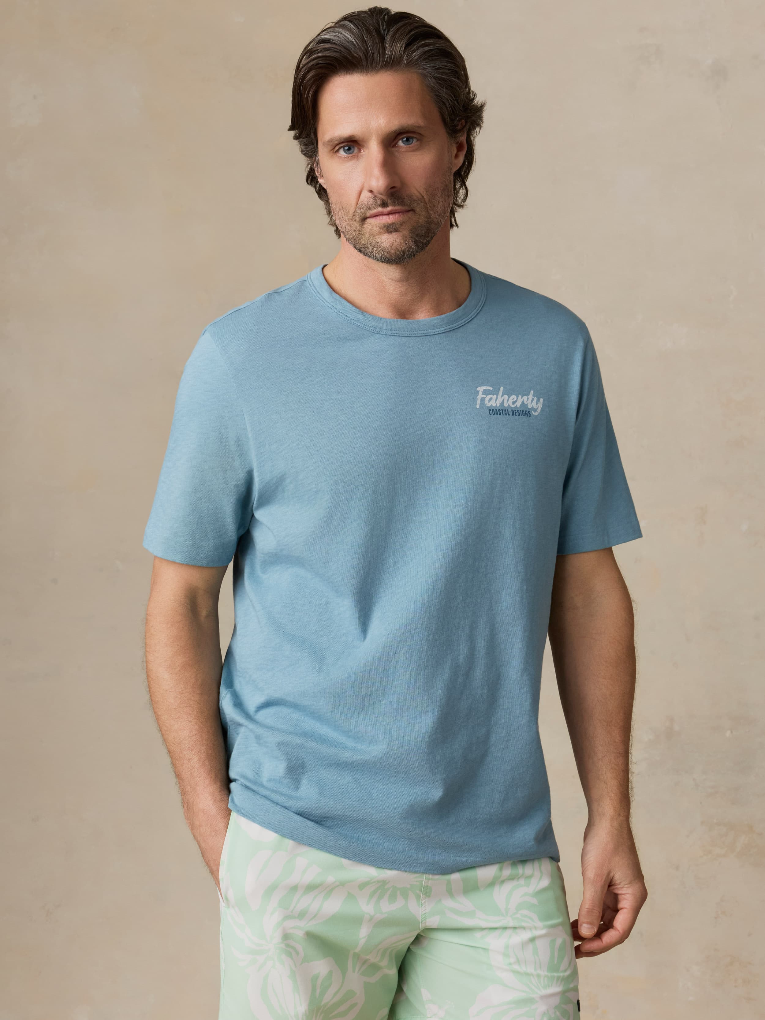 Man wearing a blue t-shirt with a Faherty logo on the left chest against a beige background