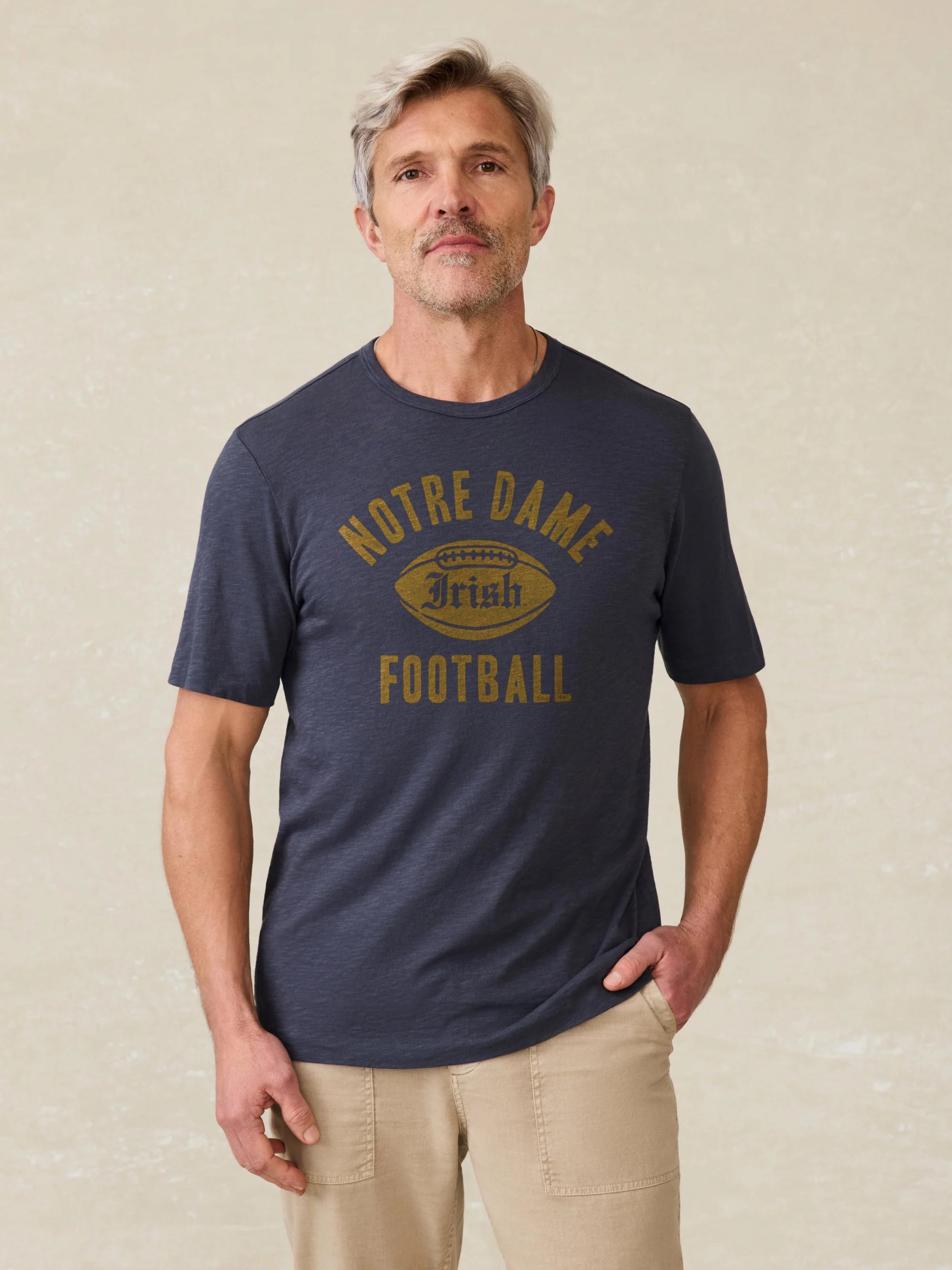 Man wearing a navy short sleeve t-shirt with Notre Dame graphic at the center front against a beige background