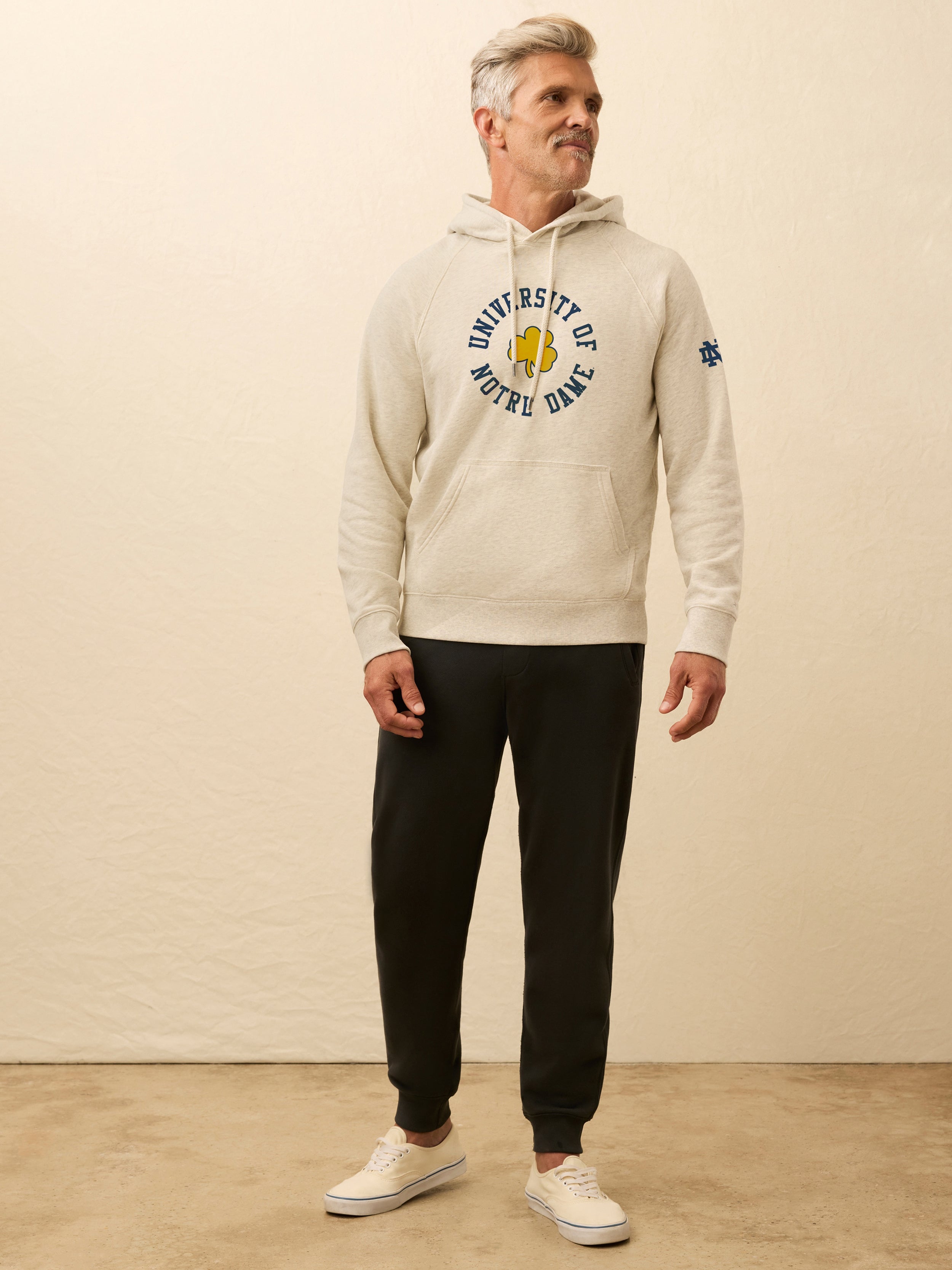 Man is wearing a heather grey hoodie with Notre Dame graphic on the center front against a beige background