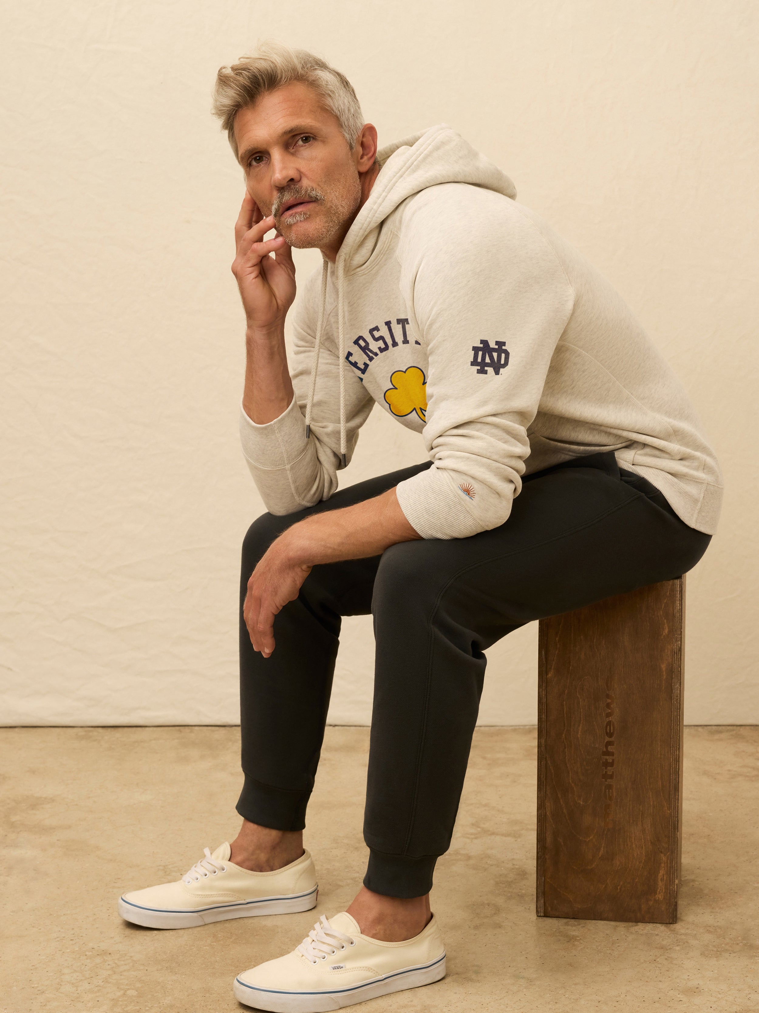 Man is wearing a heather grey hoodie with Notre Dame graphic on the center front against a beige background