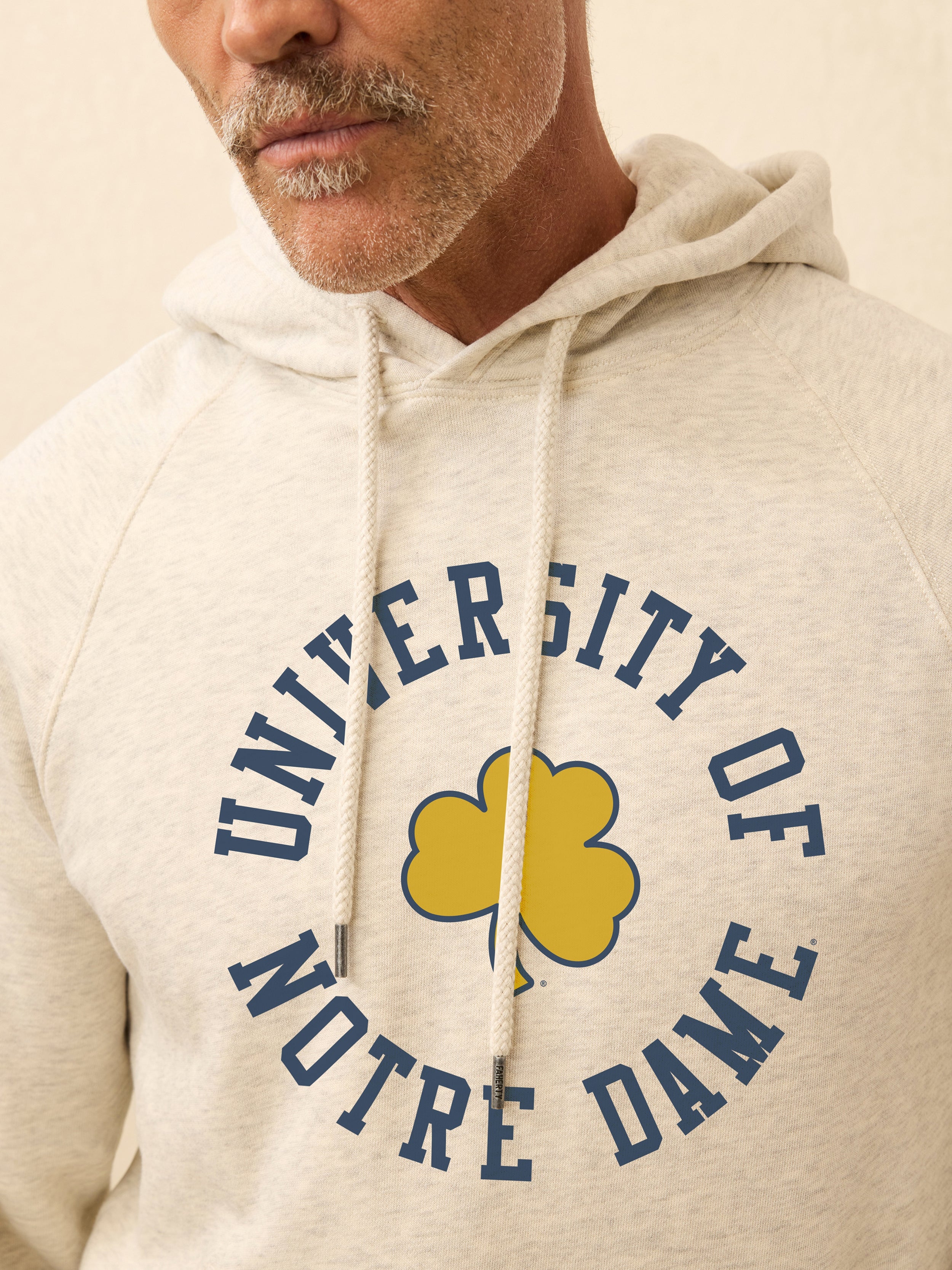 Man is wearing a heather grey hoodie with Notre Dame graphic on the center front against a beige background
