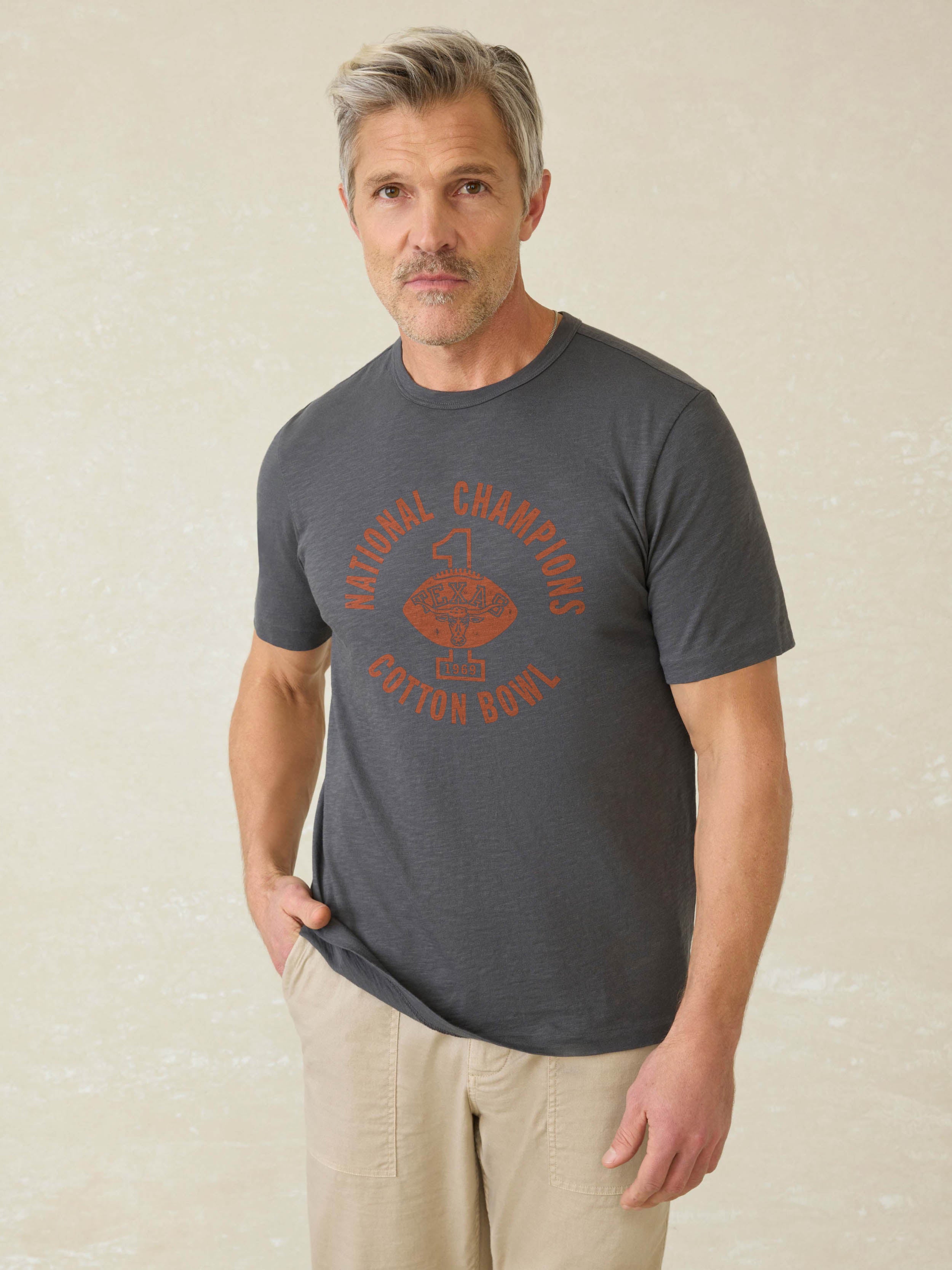 Man wearing a grey short sleeve t-shirt with UT Austin graphic on center front against a beige background