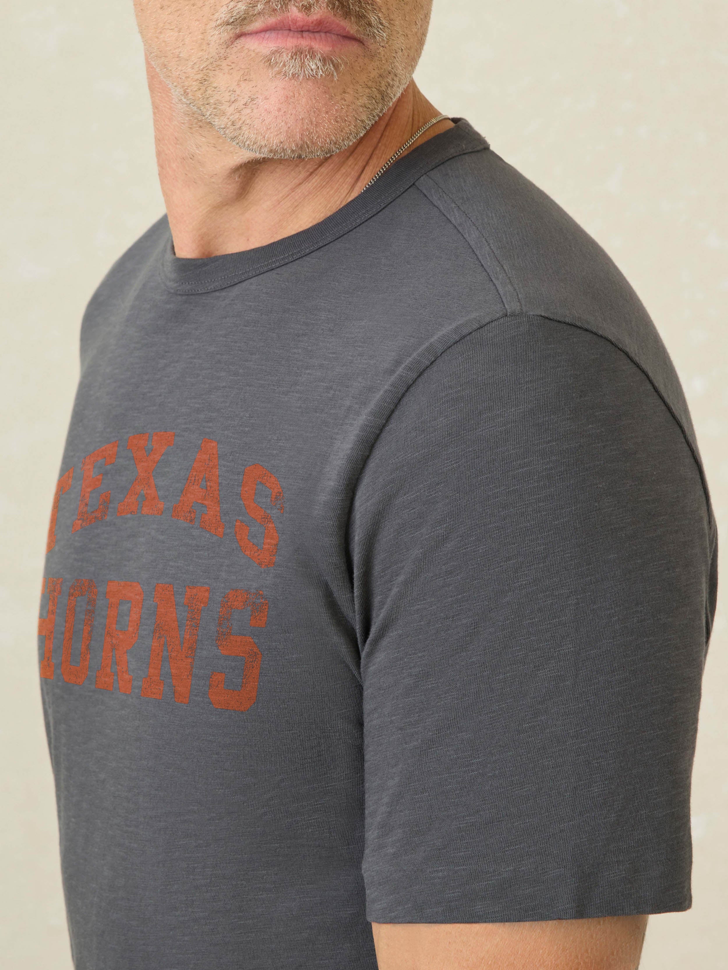 Man wearing a grey short sleeve t-shirt with UT Austin graphic logo on the center front against a beige background