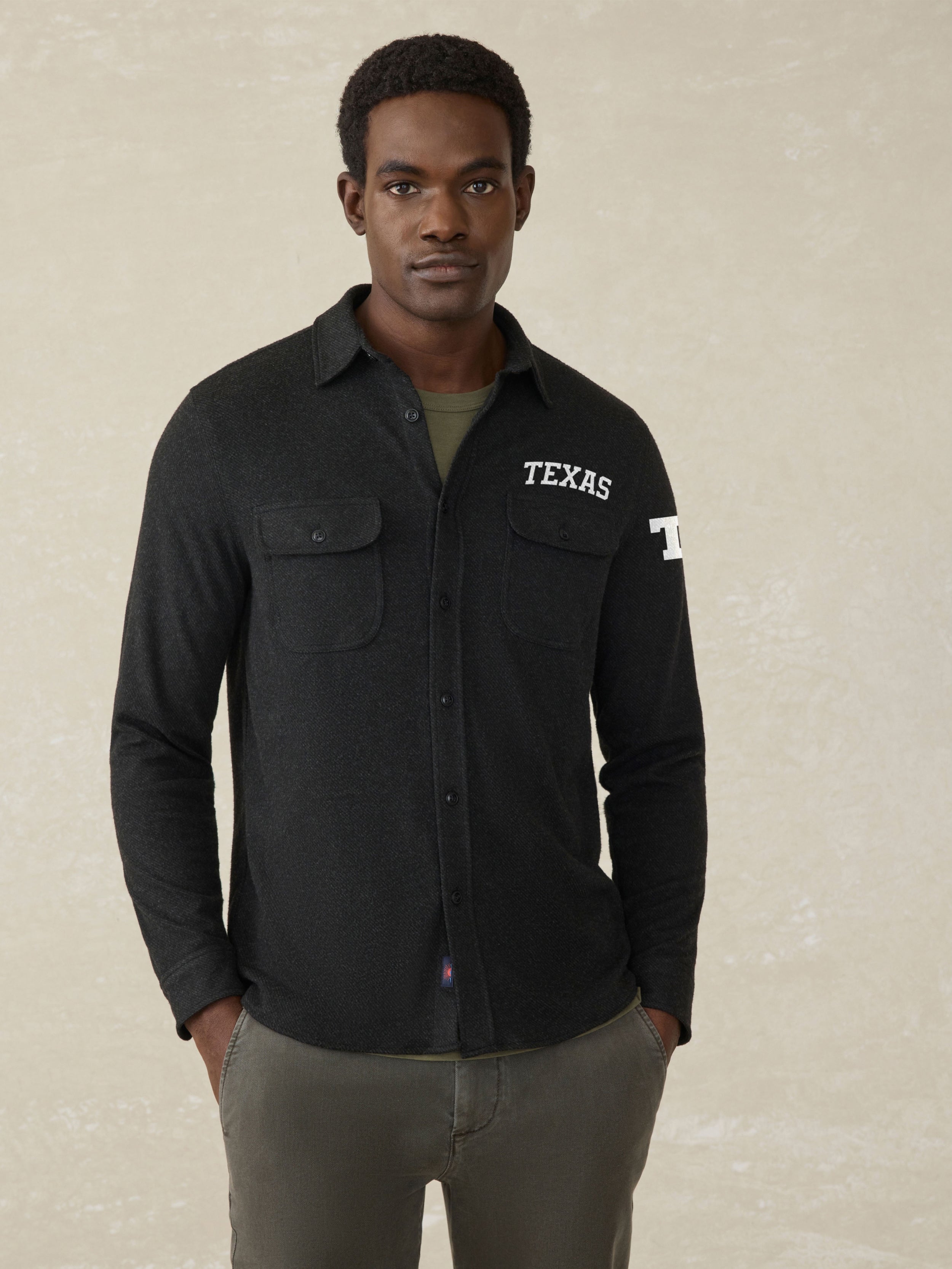 Man wearing a black long sleeve button up shirt with UT Austin graphic on the left check and shoulder against a beige background
