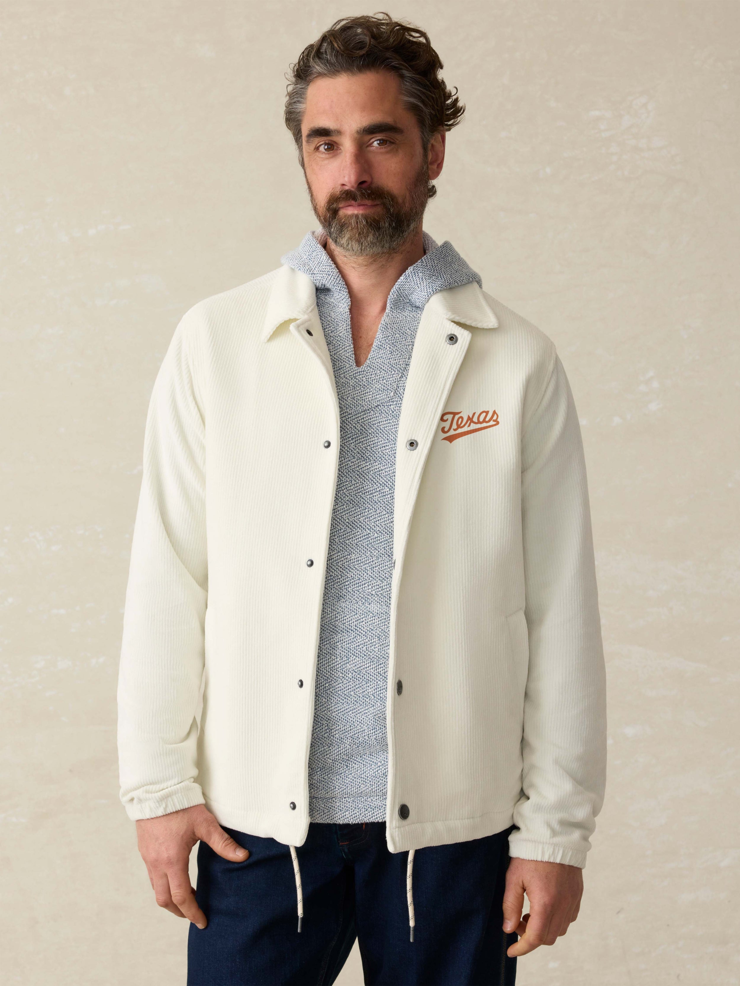 Man is wearing a white coaches jacket with UT Austin graphic on the left chest against a beige background