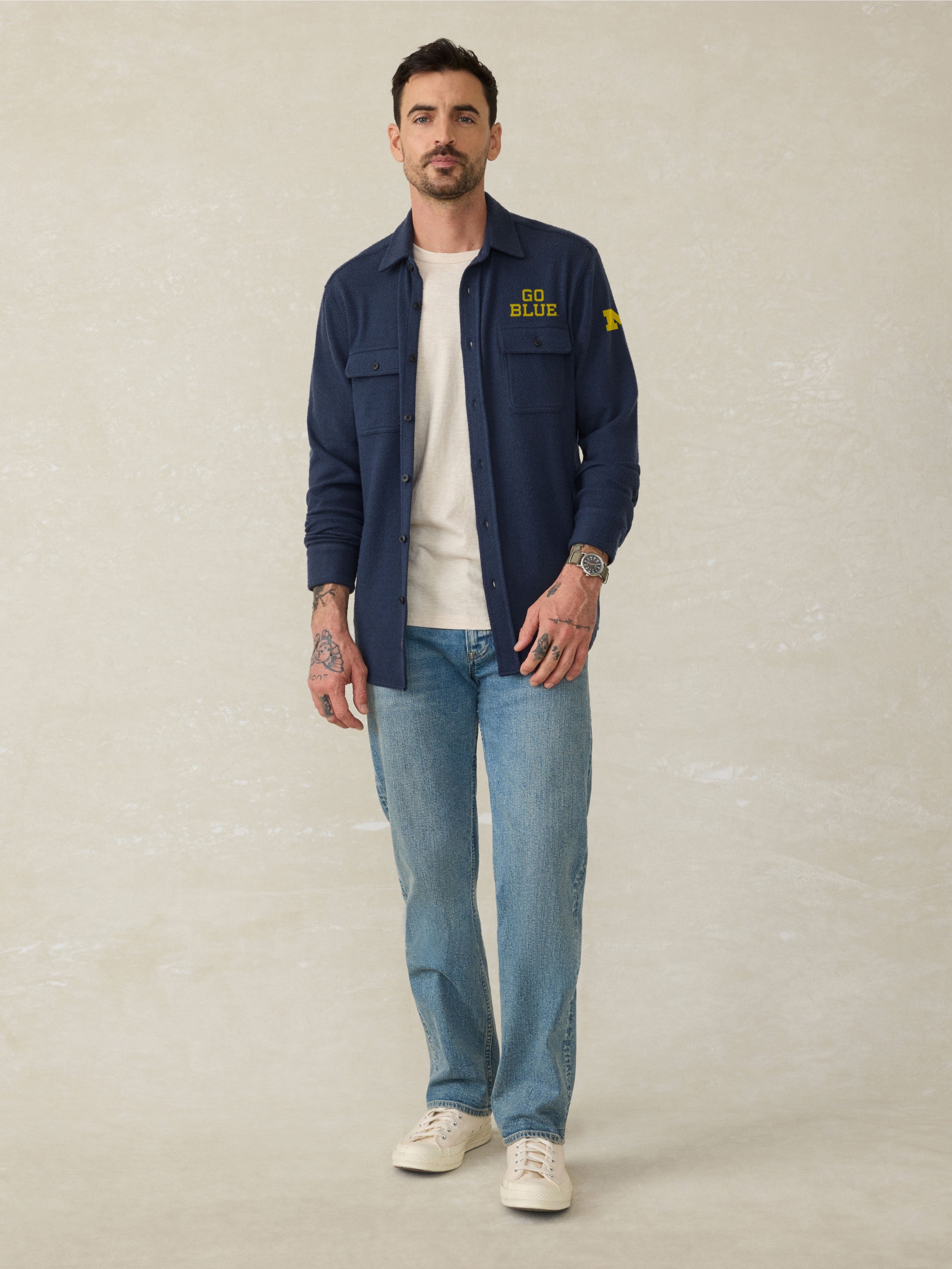 Man wearing a navy long sleeve button up shirt with Michigan graphic on left chest and left arm against a beige background