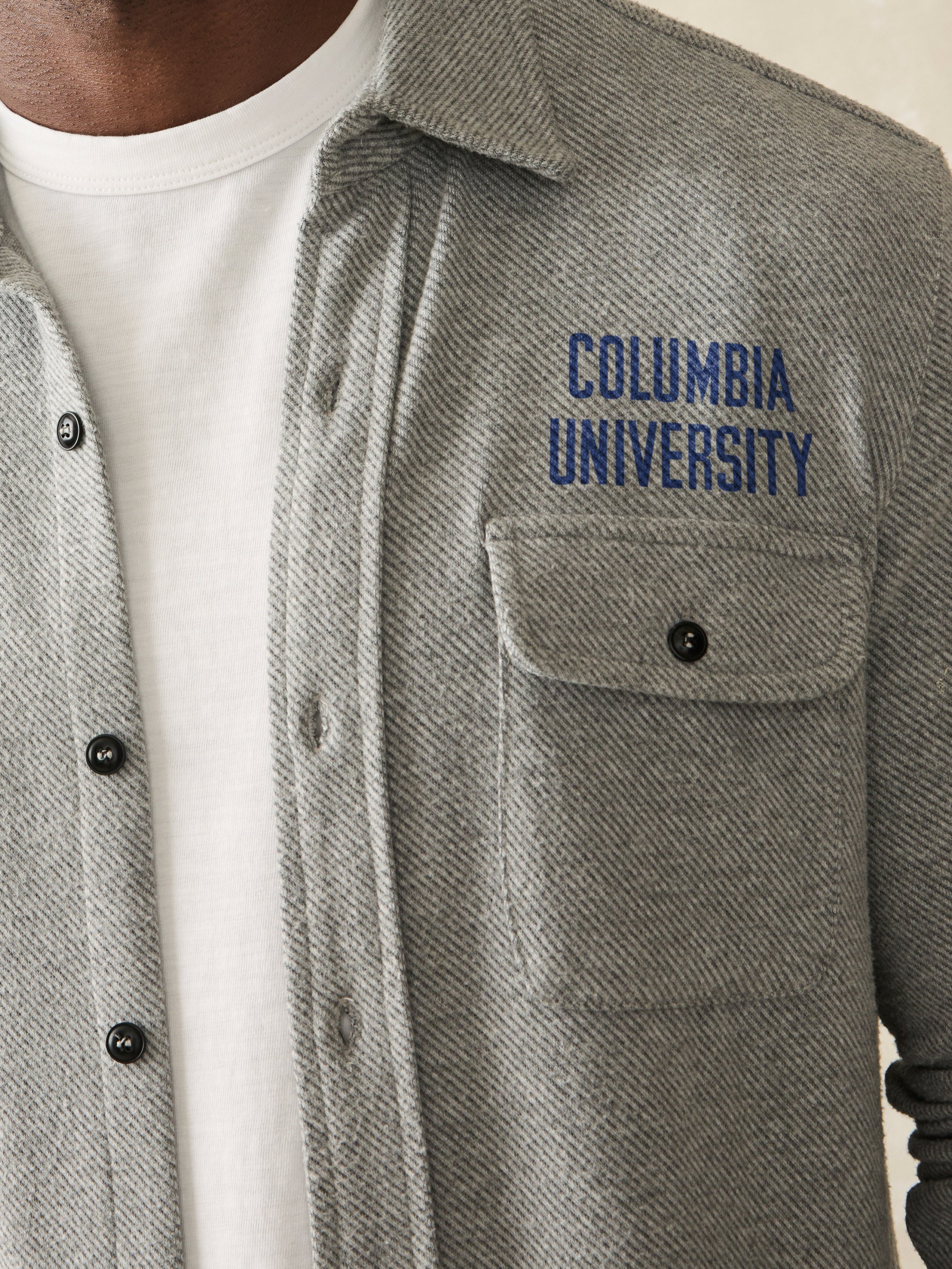 Man wearing a grey long sleeve button down shirt with Columbia University graphic on the left chest and shoulder against a beige background