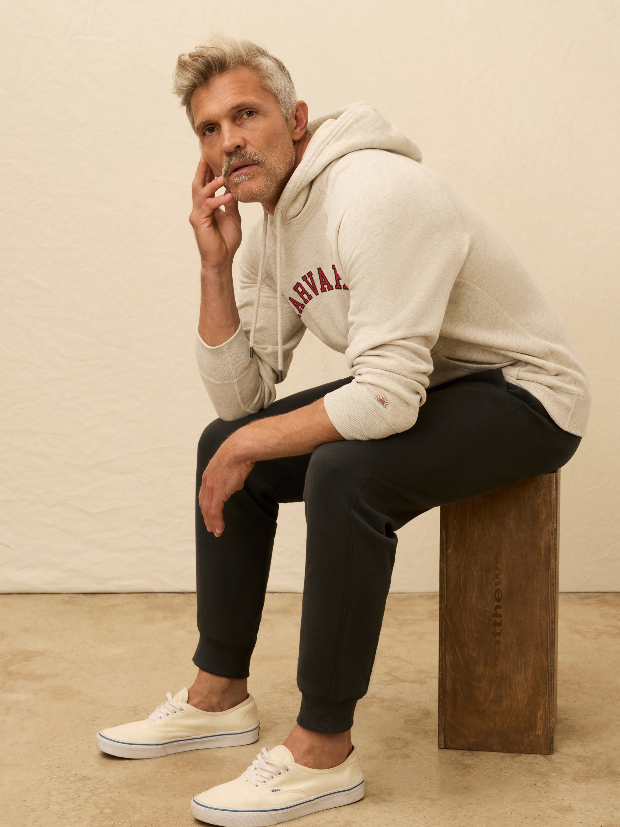 Man wearing a ivory heather hoodie with Harvard graphic on the center front against a beige background