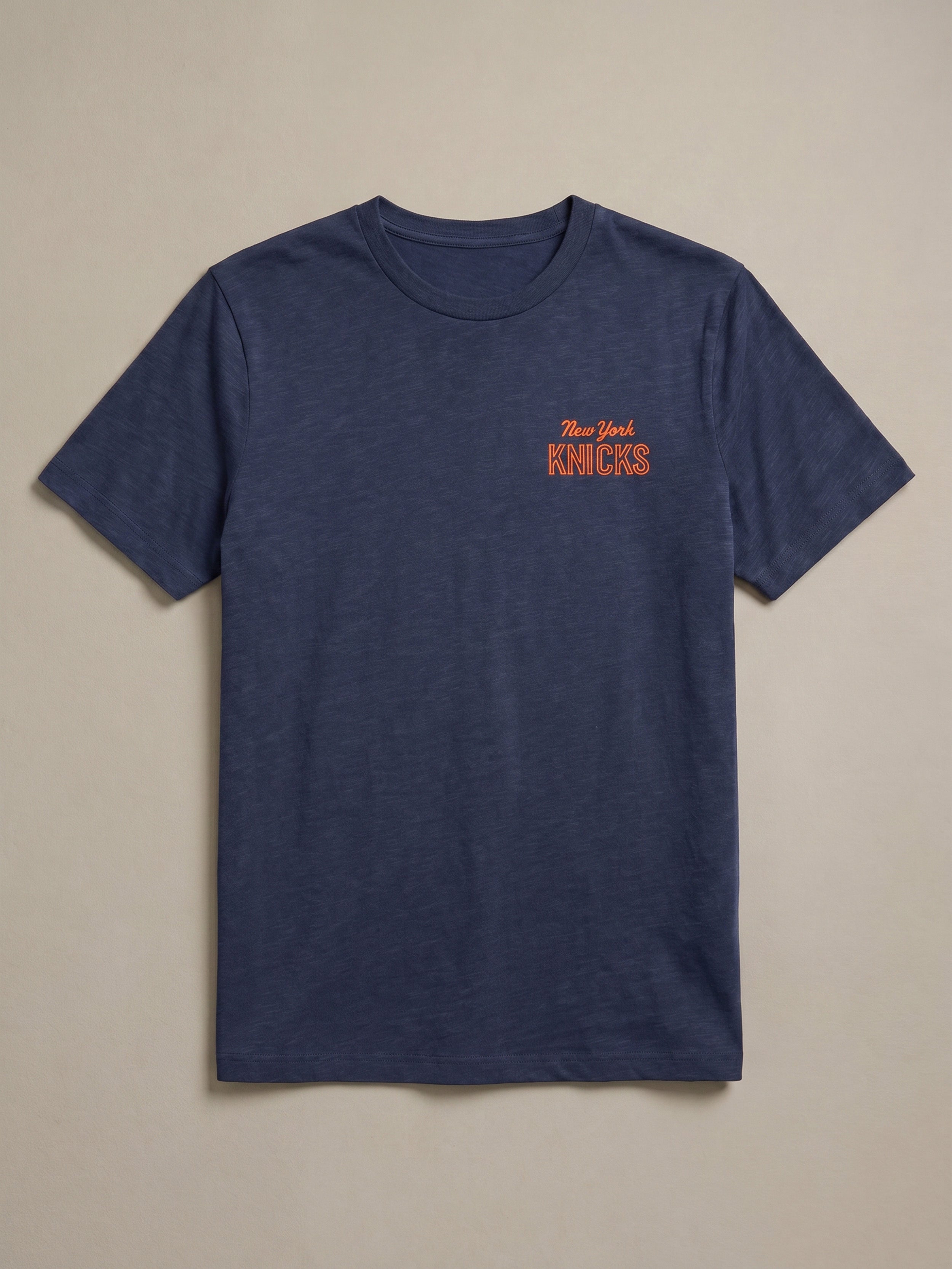 Blue t-shirt with 'New York Knicks' logo on the left chest against a beige background