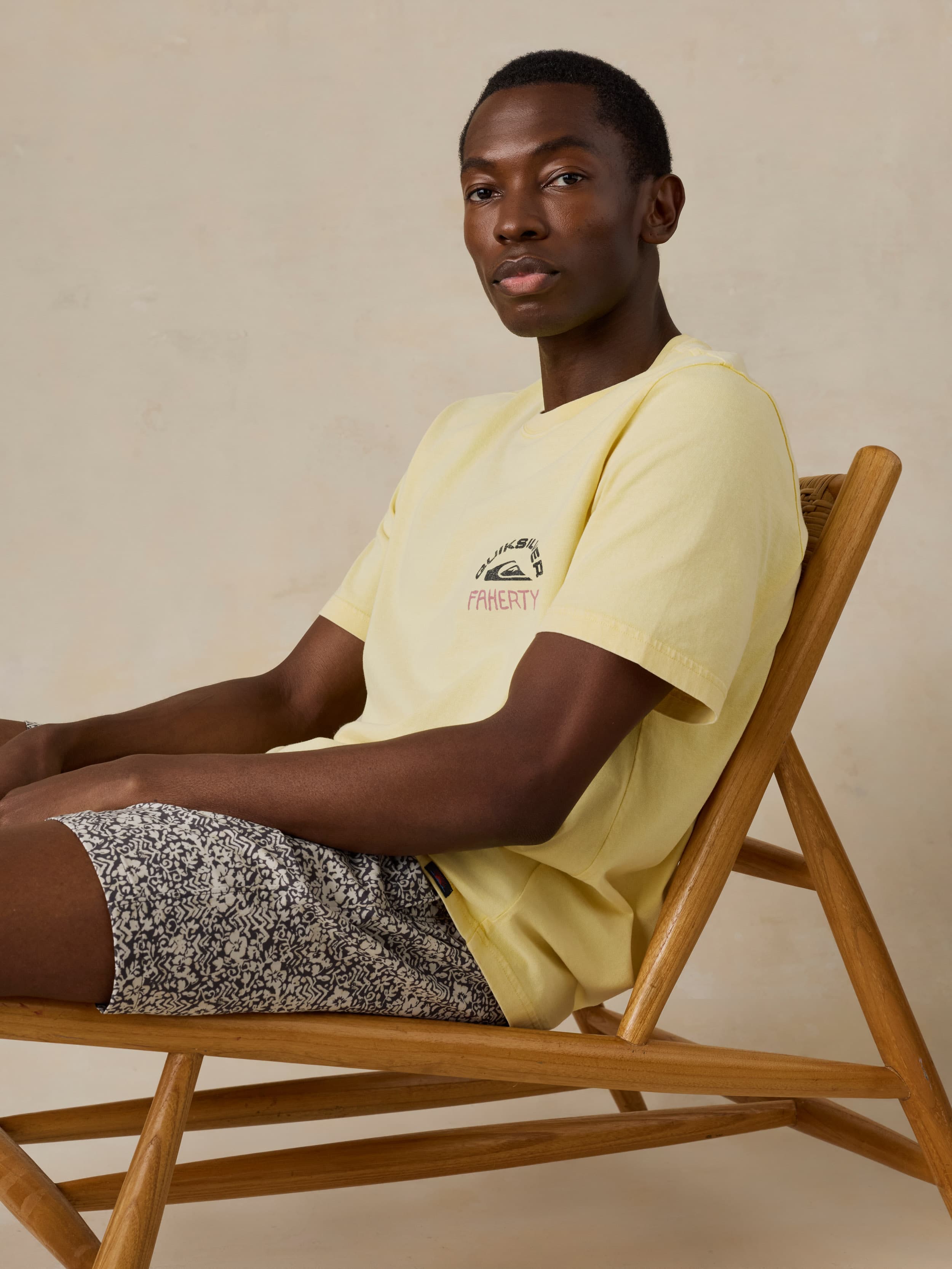 Man sitting on a wooden chair wearing a yellow t-shirt and black and white patterned shorts against a beige background