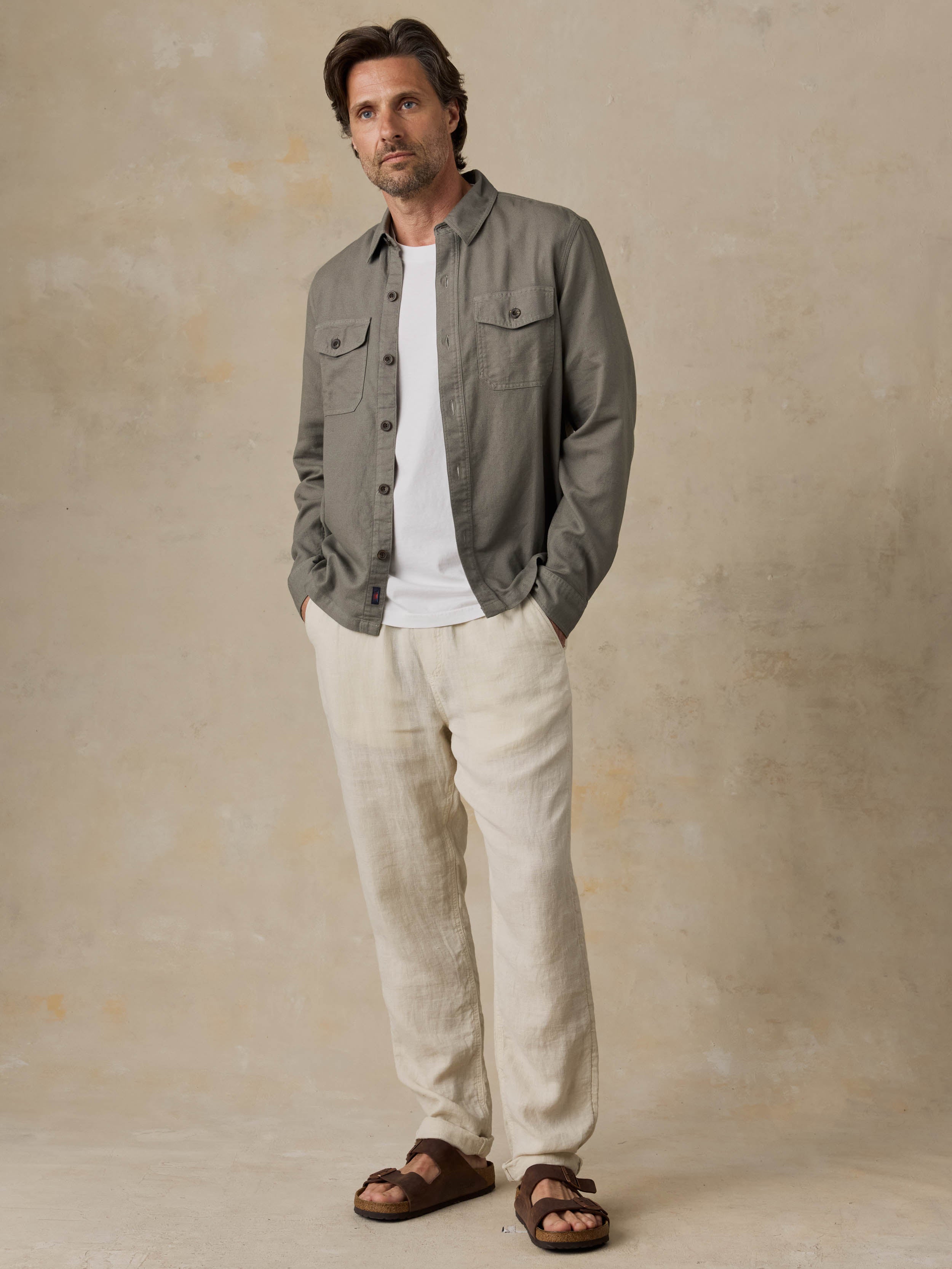 Man wearing an olive green jacket over a white shirt with beige pants against a beige background