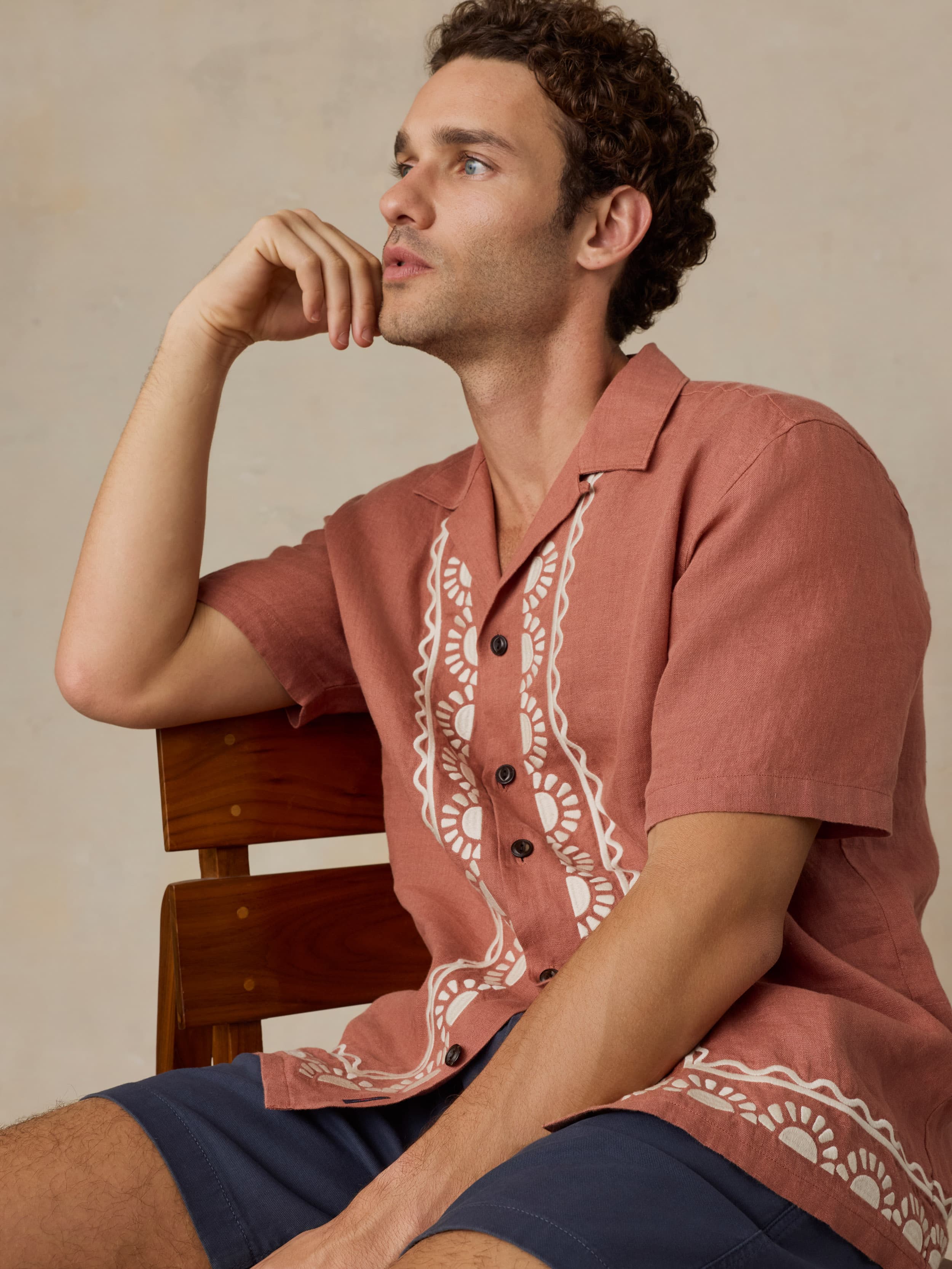 Man sitting on a wooden chair wearing a red patterned embroidered shirt and navy shorts against a beige background