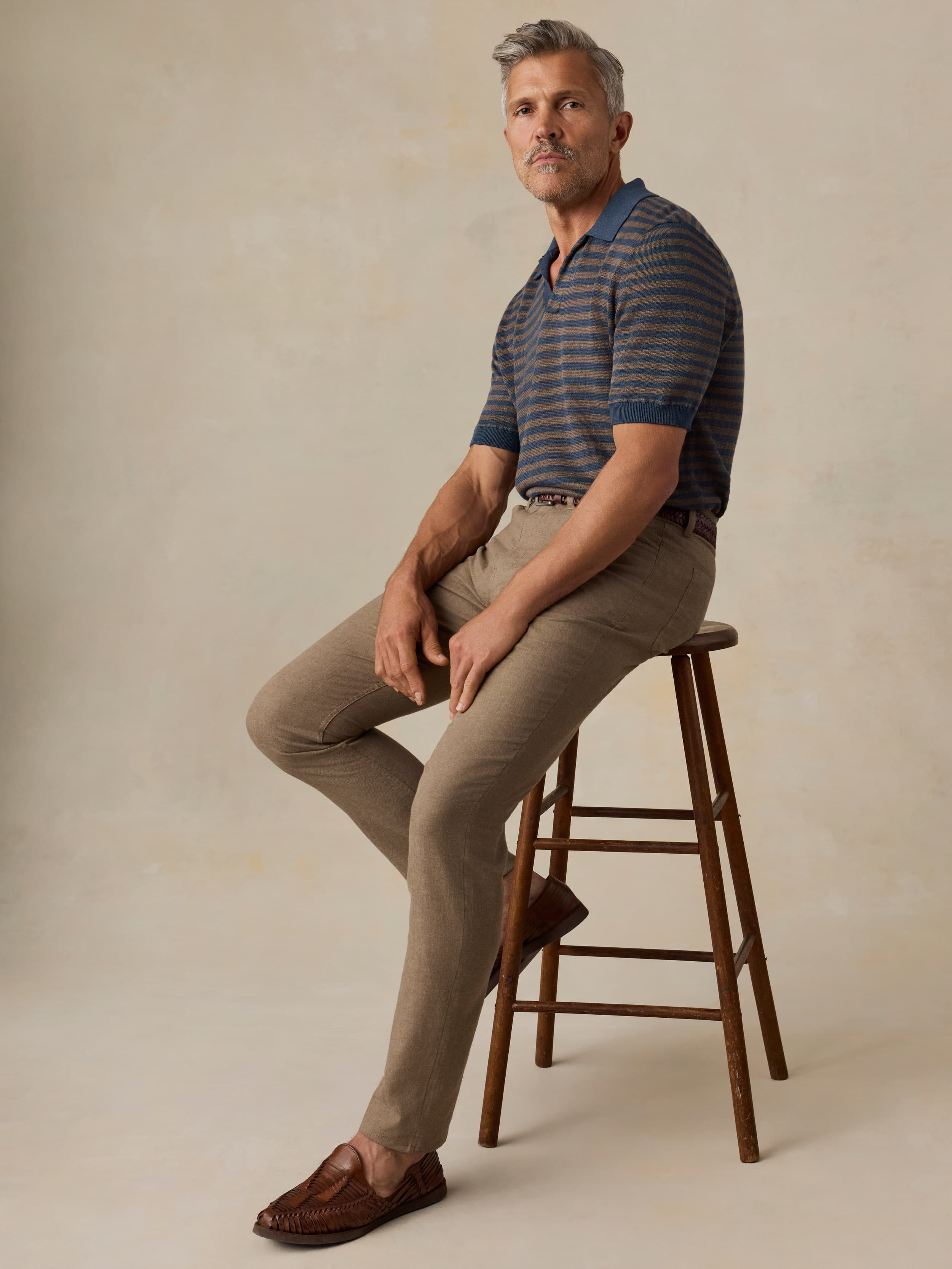 Man sitting on a wooden stool wearing a striped polo shirt and beige pants against a plain background