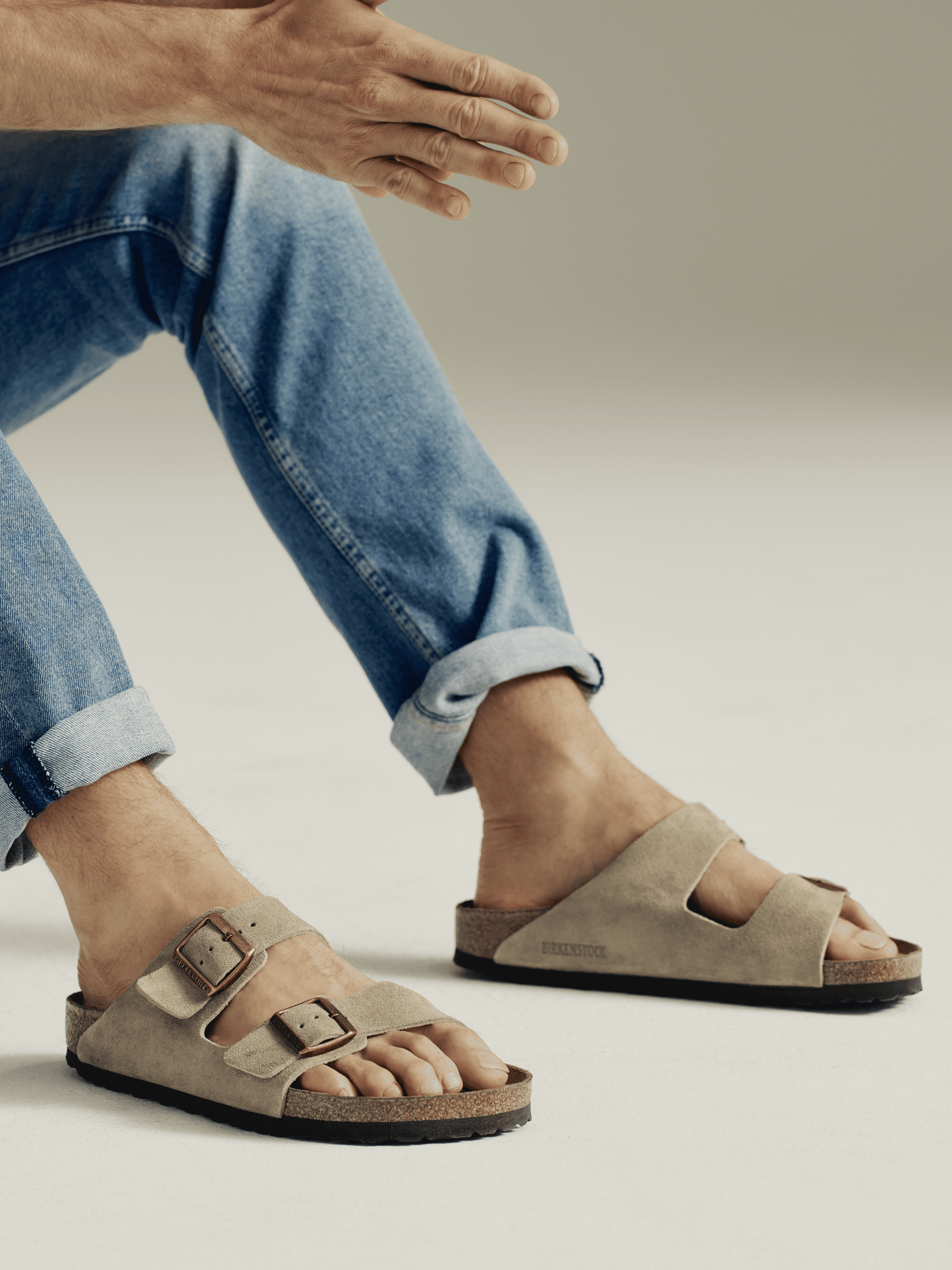 Person wearing beige suede Birkenstock sandals with rolled-up blue jeans on a neutral background