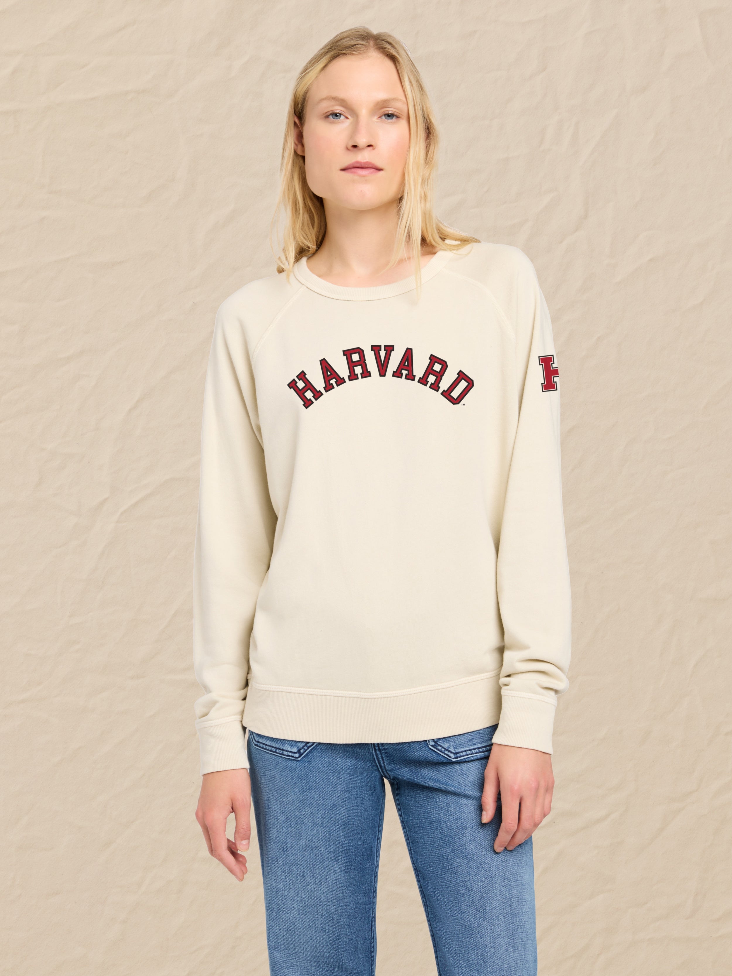 Woman wearing an ivory sweatshirt with Harvard graphic on the center front and left shoulder against a beige background