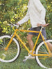 Video of a man wearing a light colored hoodie and shorts walking with a yellow bicycle in the grass